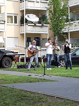 Eine kleine Band tritt im Freien vor einem Wohnhaus auf. Ein Musiker spielt Gitarre und singt, während andere Trompete spielen. Im Hintergrund stehen Autos und ein Lautsprecher.