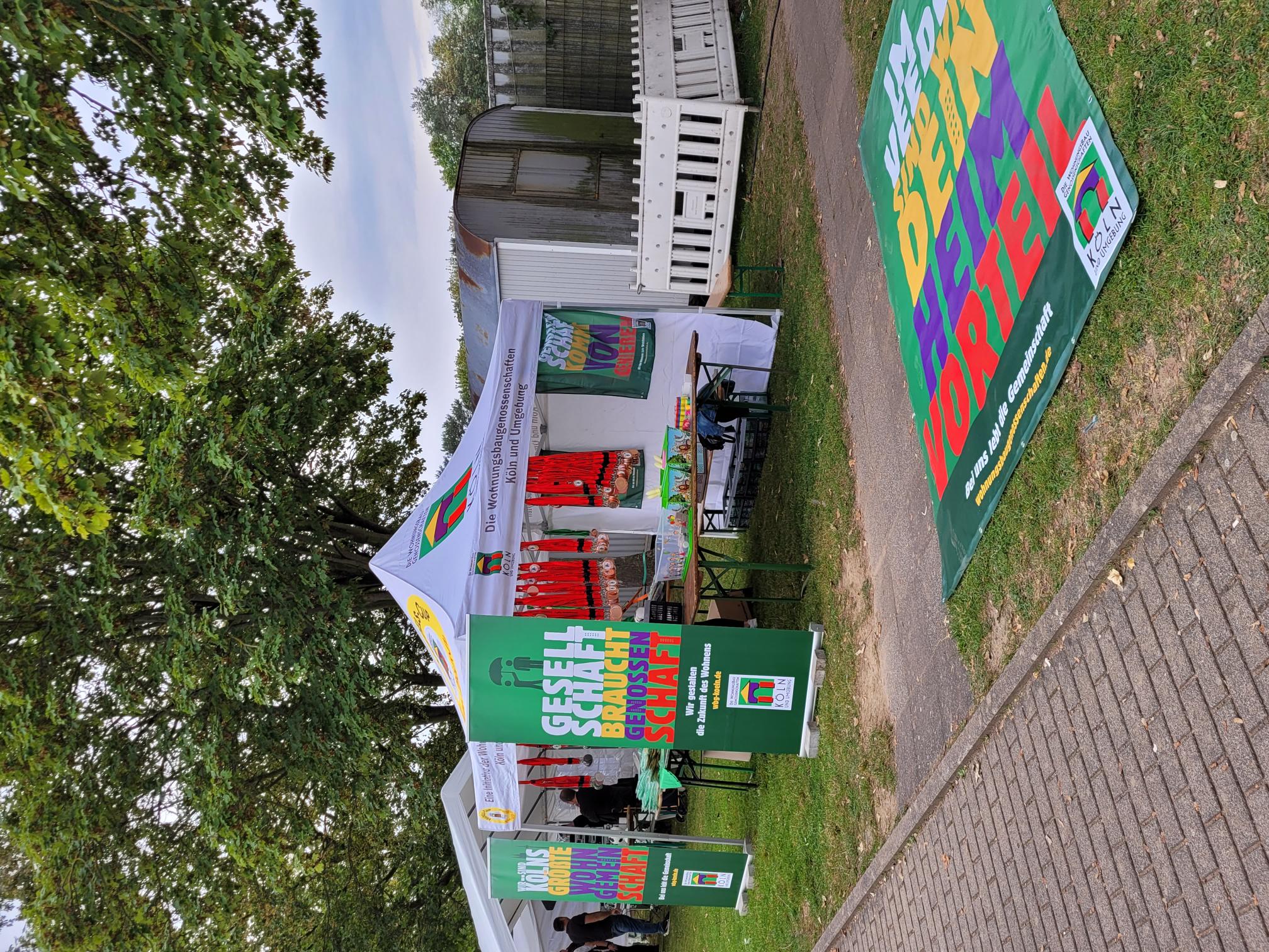 Ein weißer Infostand der Wohnungsbaugenossenschaften mit Bannern, einem Tisch voller Medaillen und Werbeartikeln, umgeben von großen Bäumen.