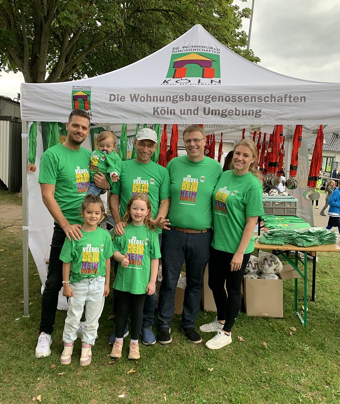 Eine fröhliche Gruppe aus Erwachsenen und Kindern in grünen T-Shirts posiert vor dem Stand der Wohnungsbaugenossenschaften Köln und Umgebung.