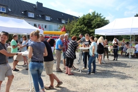 Ein geselliges Fest bei sonnigem Wetter, bei dem Menschen an Stehtischen unter weißen Pavillons zusammenkommen. Im Hintergrund sind eine Hüpfburg und Wohnhäuser zu sehen. Die entspannte Atmosphäre lädt zum Austausch und Verweilen ein.