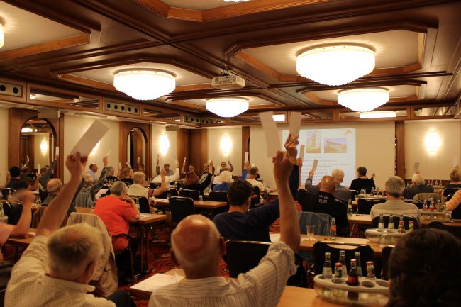 Eine Versammlung in einem eleganten Konferenzraum mit Kronleuchtern, bei der die Teilnehmenden mit erhobenen Stimmzetteln über ein Thema abstimmen. Im Hintergrund ist eine Präsentation auf einer Leinwand zu sehen.