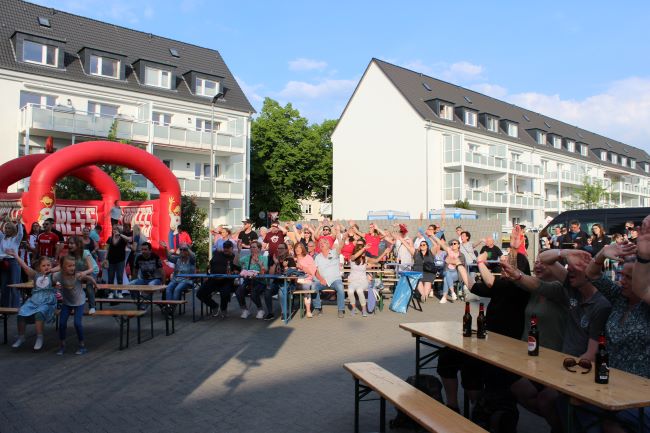 Eine große Menschenmenge sitzt und steht an Biertischen bei einer Veranstaltung im Freien, mit aufgeregten Gesten und Blicken. Im Hintergrund sind Wohngebäude und eine rote Hüpfburg zu sehen.