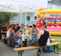 Eine fröhliche Sommerfest-Szene, bei der Familien und Freunde an Biertischen unter einem Pavillon zusammensitzen. Im Hintergrund spielen Kinder ausgelassen auf einer bunten Hüpfburg mit einem lachenden Clown-Gesicht. Die entspannte Atmosphäre lädt zum gemeinsamen Verweilen ein.