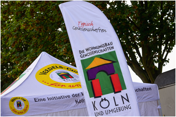 Ein Informationsstand der Wohnungsbaugenossenschaften Köln und Umgebung mit einem weißen Pavillon und einer großen Werbefahne. Beide sind mit dem Logo und dem Schriftzug des Veedels-Cups versehen. Im Hintergrund ragen grüne Baumkronen in den Himmel.
